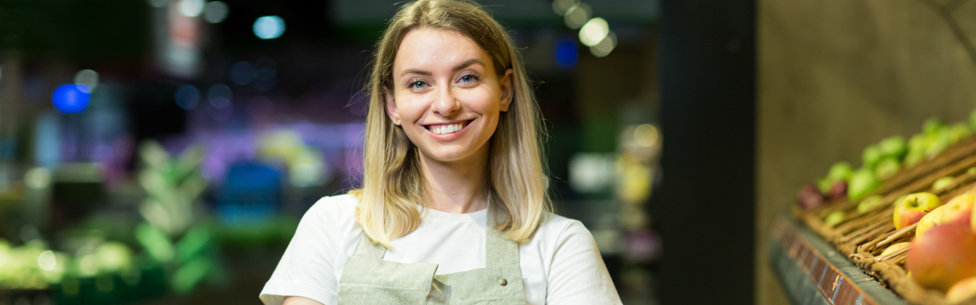 Freundliche Supermarktmitarbeiterin mit Schürze steht lächelnd in der Obst- und Gemüseabteilung